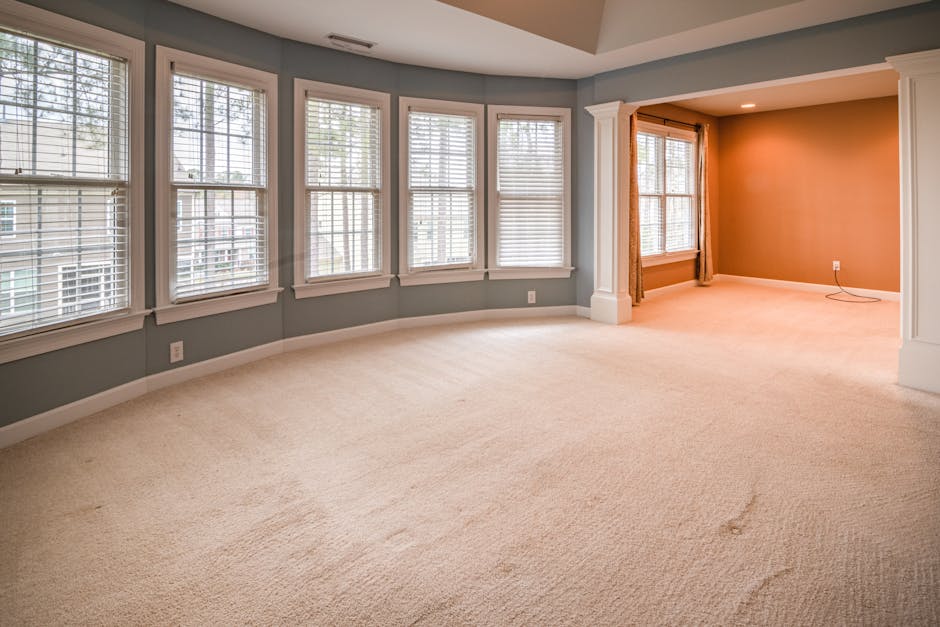 A professional cleaner dressed in a full white protective suit, face mask, and gloves is performing surface cleaning on a beige carpet in a bright, modern living room. The cleaner is using a steam or 