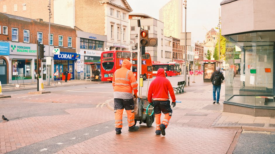Two professional cleaners from Harold Hill Carpet Cleaners are performing surface cleaning and deep cleaning tasks on the pavement of a busy urban street. They are wearing high-visibility orange and r