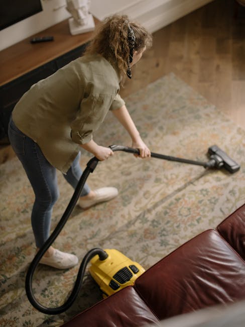 A professional cleaner from Harold Hill Carpet Cleaners dressed in a white protective suit, face mask, gloves, and blue shoe covers, is performing deep cleaning on a beige carpet in a bright, modern l