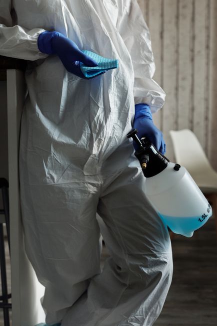 A person dressed in full white protective cleaning gear, including a hood, mask, gloves, and goggles, is performing surface cleaning in a modern living room. They are leaning over a black round table,